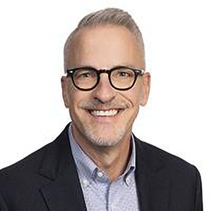 The image shows a middle-aged man with short, gray hair and a neatly trimmed beard. He is wearing black-rimmed glasses and a light blue collared shirt under a dark blazer. The man is smiling, and the background is plain white.